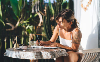 A woman with a tattoo on her arm sits at a table outdoors, writing in a notebook. She is surrounded by greenery and appears focused. The table holds a bell and other small items. The setting is relaxed and sunny.