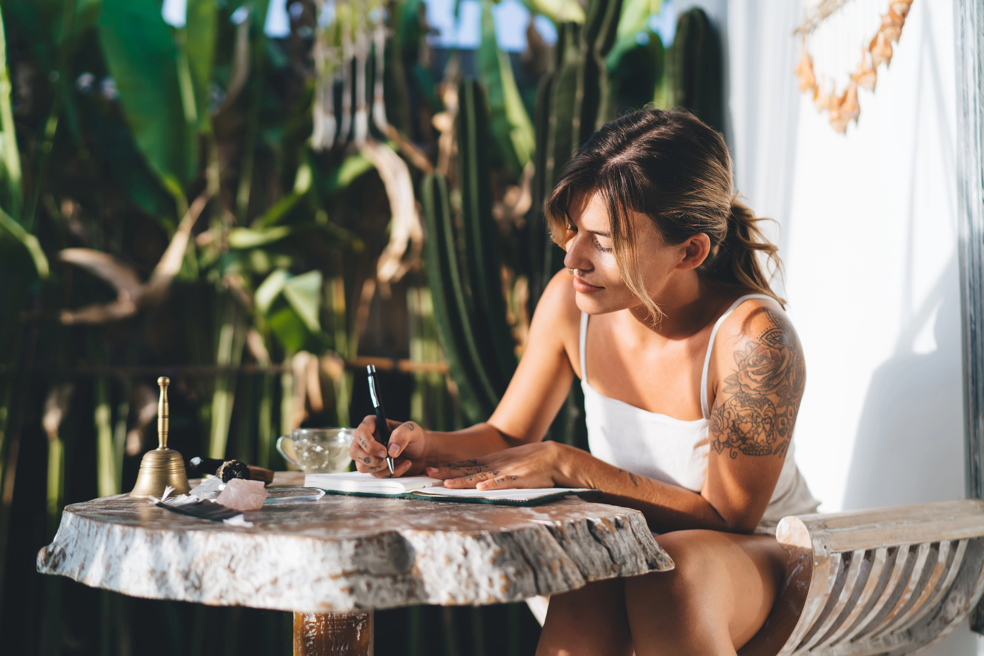 A woman with a tattoo on her arm sits at a table outdoors, writing in a notebook. She is surrounded by greenery and appears focused. The table holds a bell and other small items. The setting is relaxed and sunny.