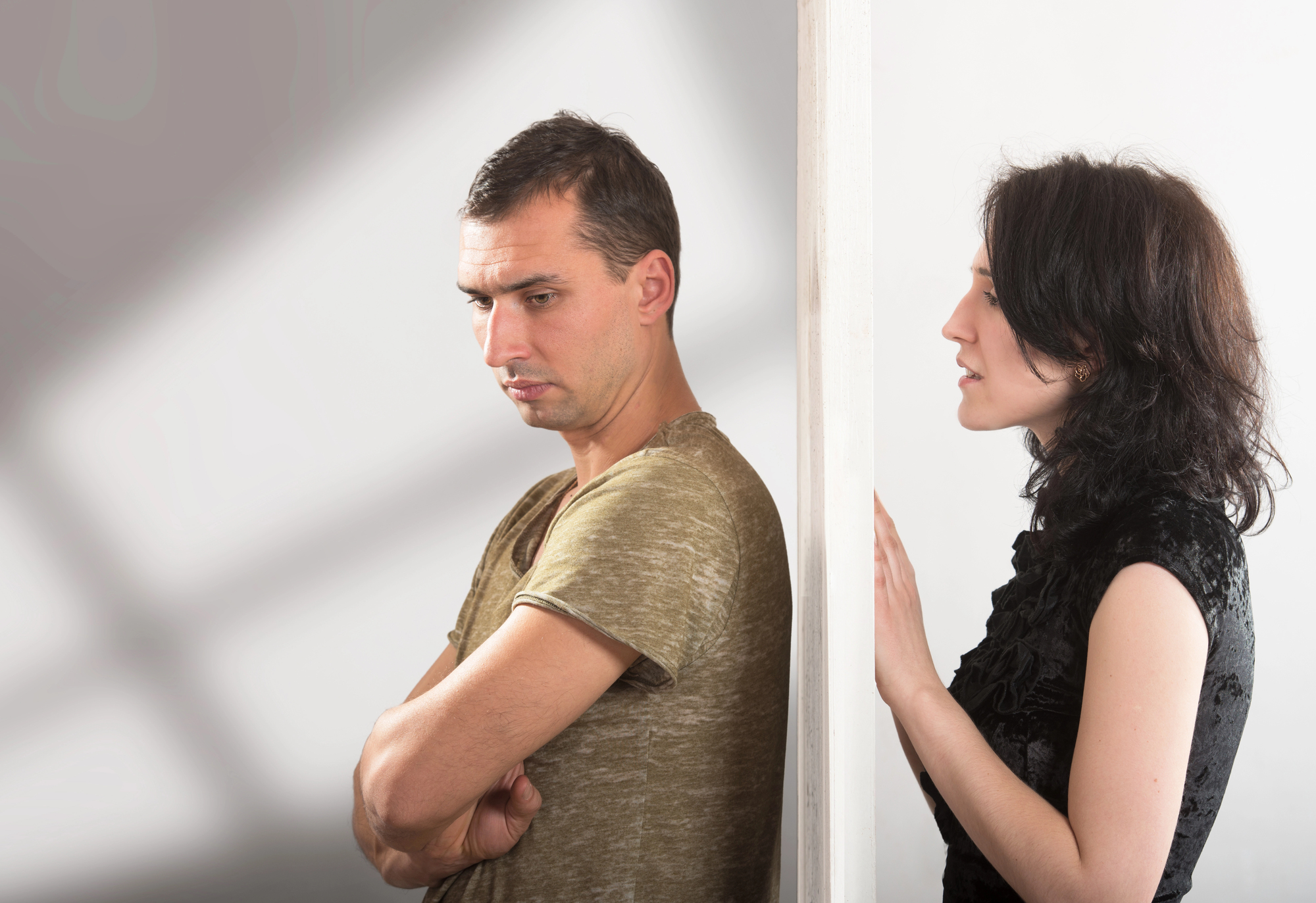 A man and a woman are standing back to back, separated by a wall. The man has his arms crossed and looks away, while the woman appears to be talking or pleading. The image suggests tension or a disagreement between them.