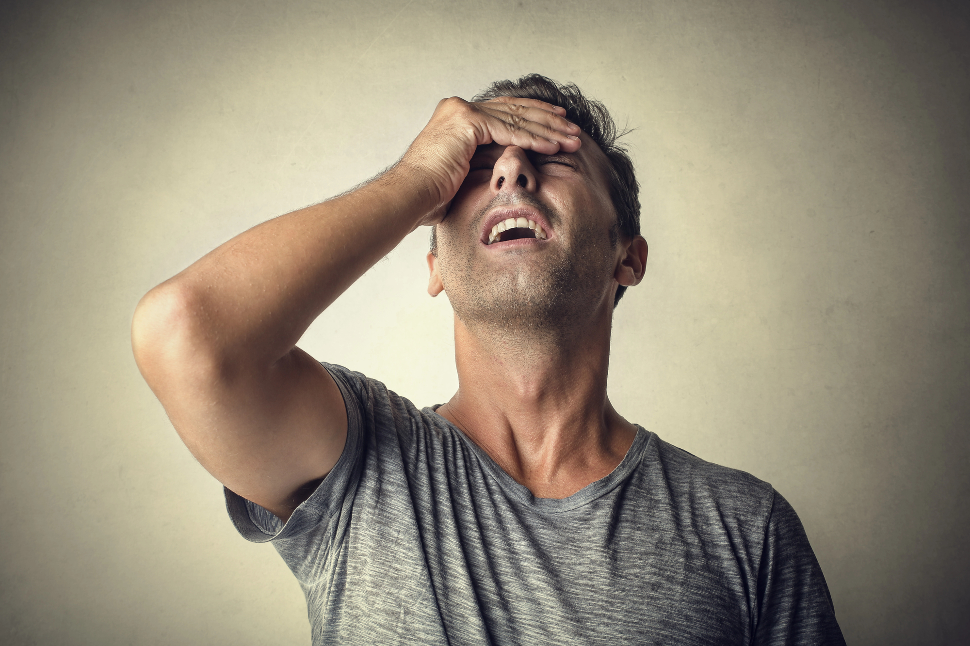 A man in a gray t-shirt stands against a plain background, looking up with his eyes closed. He is holding his forehead with one hand, appearing frustrated or stressed.