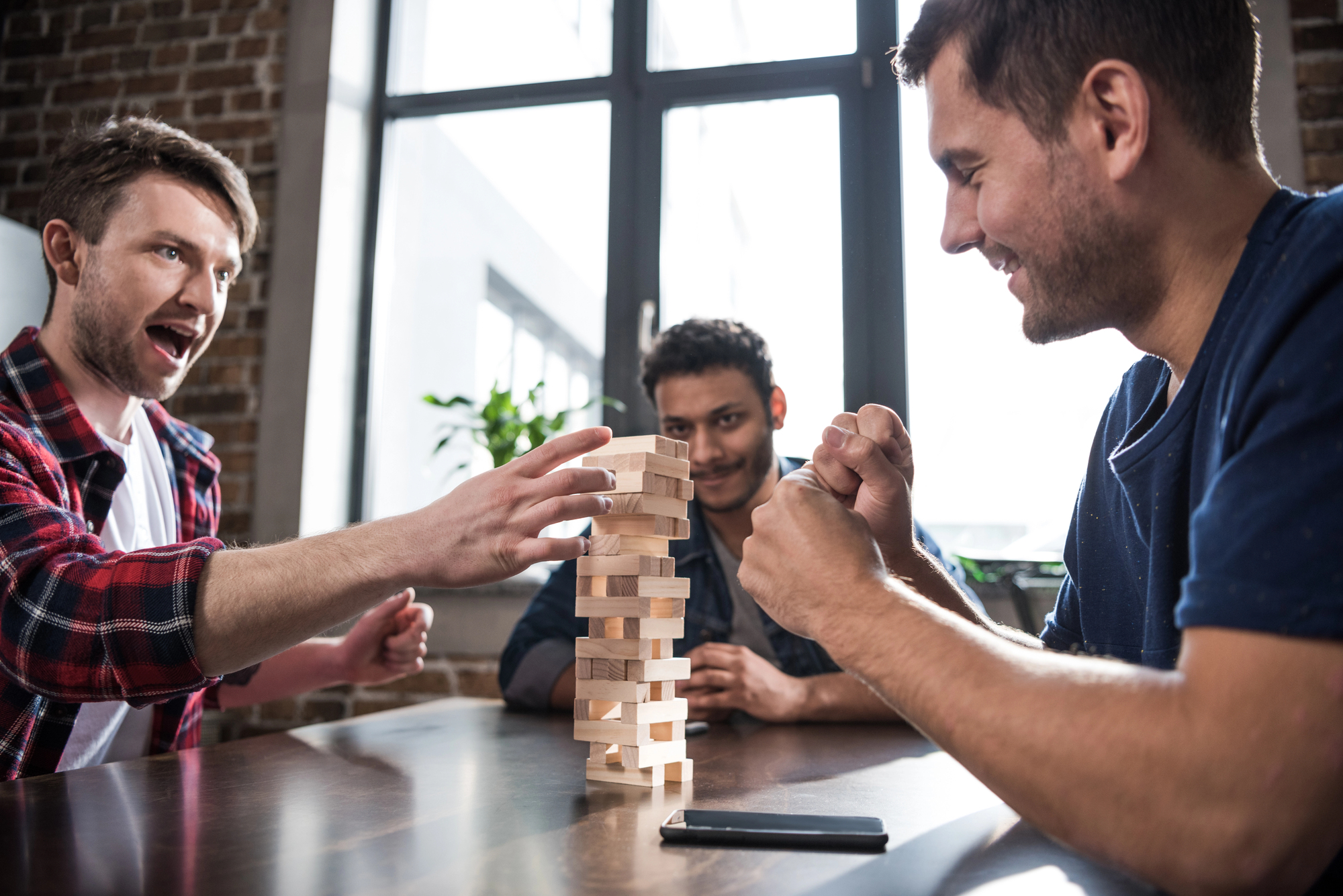 Three men are playing Jenga at a wooden table. One man is carefully removing a block while the others watch intently. They are in a bright room with large windows and a brick wall. A smartphone is on the table near them.