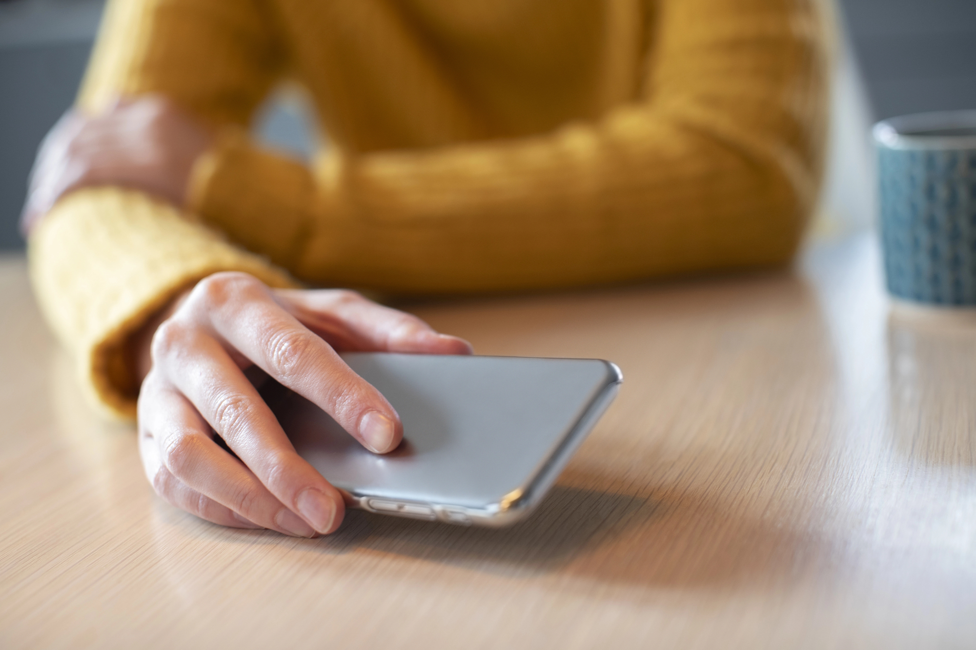 A person in a yellow sweater sits at a table, gently resting their hand on a smartphone. A coffee mug is partially visible in the background.