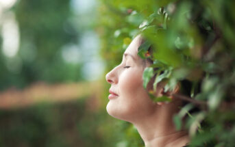 A woman with closed eyes leans against green foliage, appearing relaxed and content. The background is softly blurred, highlighting a serene, nature-focused atmosphere.