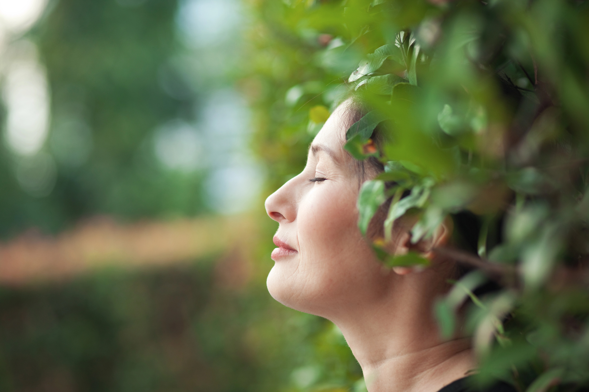 A woman with closed eyes leans against green foliage, appearing relaxed and content. The background is softly blurred, highlighting a serene, nature-focused atmosphere.