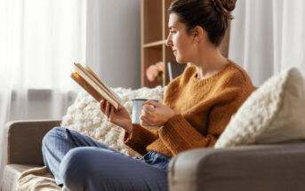 A woman sits on a couch, wearing a brown sweater and holding a book and a mug. She has her hair in a bun and is looking towards the window. The room is softly lit, with a cozy blanket and a shelf in the background.