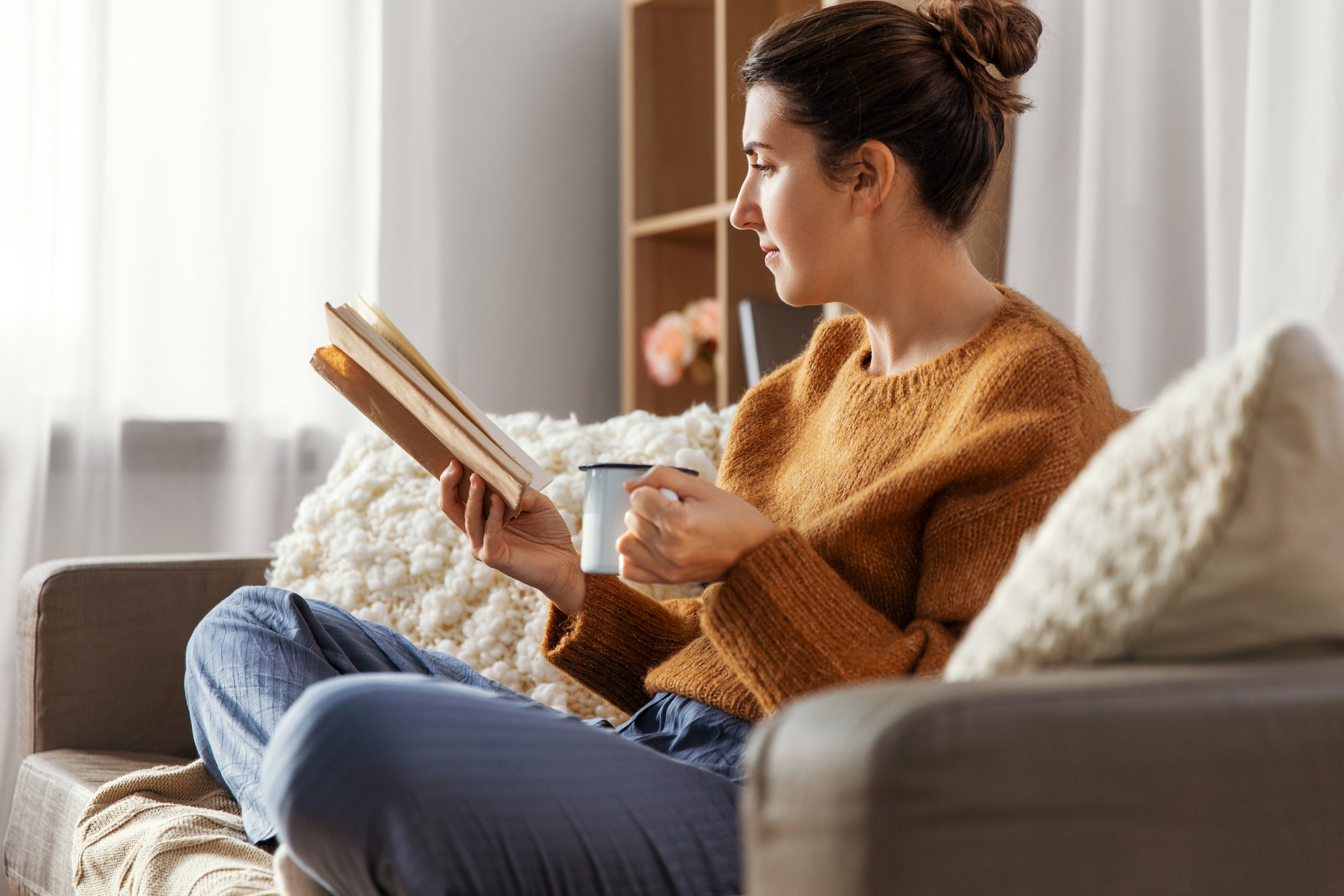 A woman sits on a couch, wearing a brown sweater and holding a book and a mug. She has her hair in a bun and is looking towards the window. The room is softly lit, with a cozy blanket and a shelf in the background.