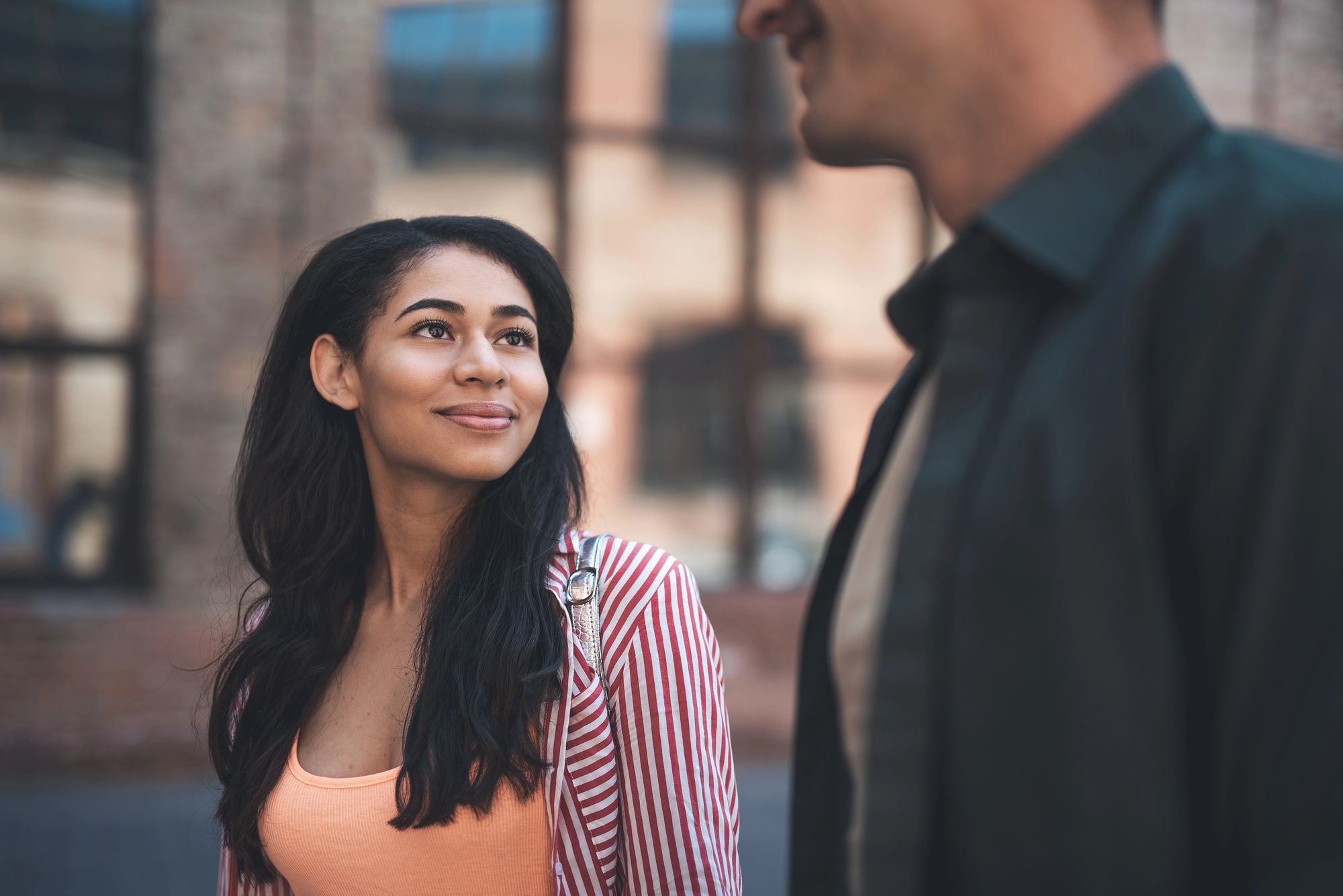 A woman with long dark hair, wearing a striped jacket and orange top, smiles while looking at a man in a dark shirt. They are standing outdoors in front of a building with large windows.