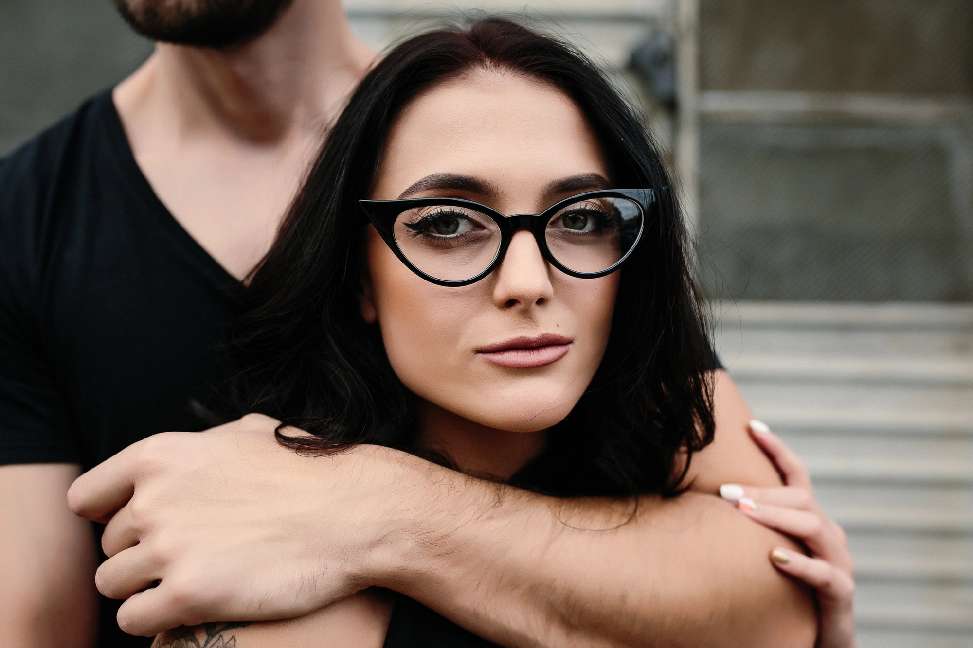 A woman with long dark hair and cat-eye glasses is looking at the camera. She is being gently embraced from behind by a person in a black shirt, whose arms are wrapped around her shoulders.