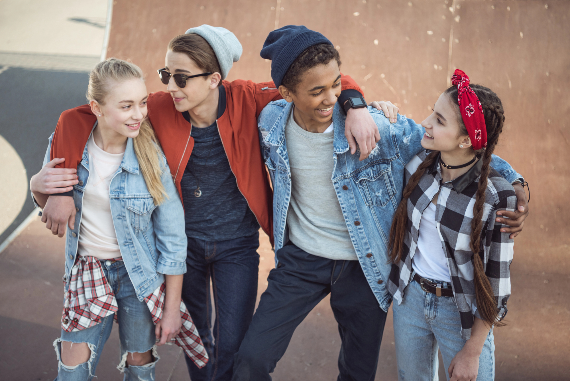 Four teenagers at a skatepark, with arms around each other, smiling and dressed in casual clothes. Two are wearing denim jackets, one in a red jacket, and the girl on the right in a plaid shirt. Skate ramp visible in the background.