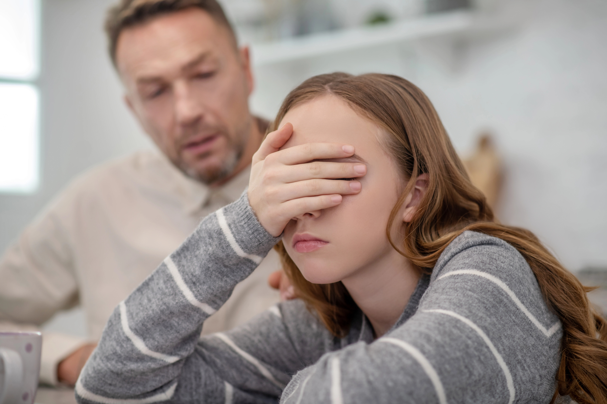 A teenage girl sits at a table, covering her eyes with her hand, appearing upset. A man sits nearby, looking at her with concern, in a cozy room setting.