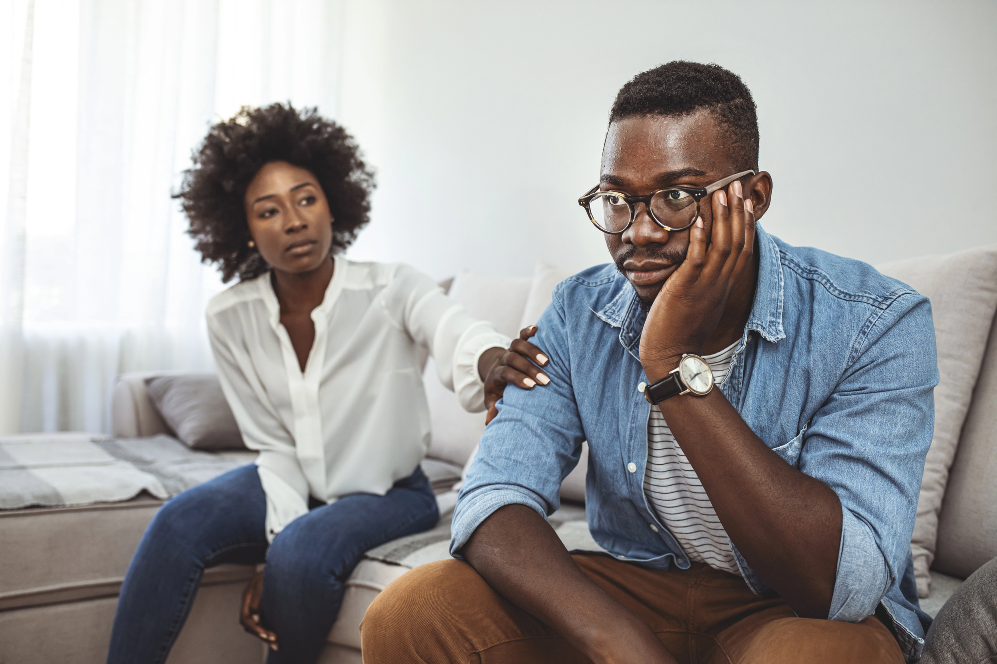 A woman in a white blouse gently touches the shoulder of a man in glasses and a denim shirt, who appears upset. They are seated on a couch in a well-lit room, suggesting a supportive or comforting moment.