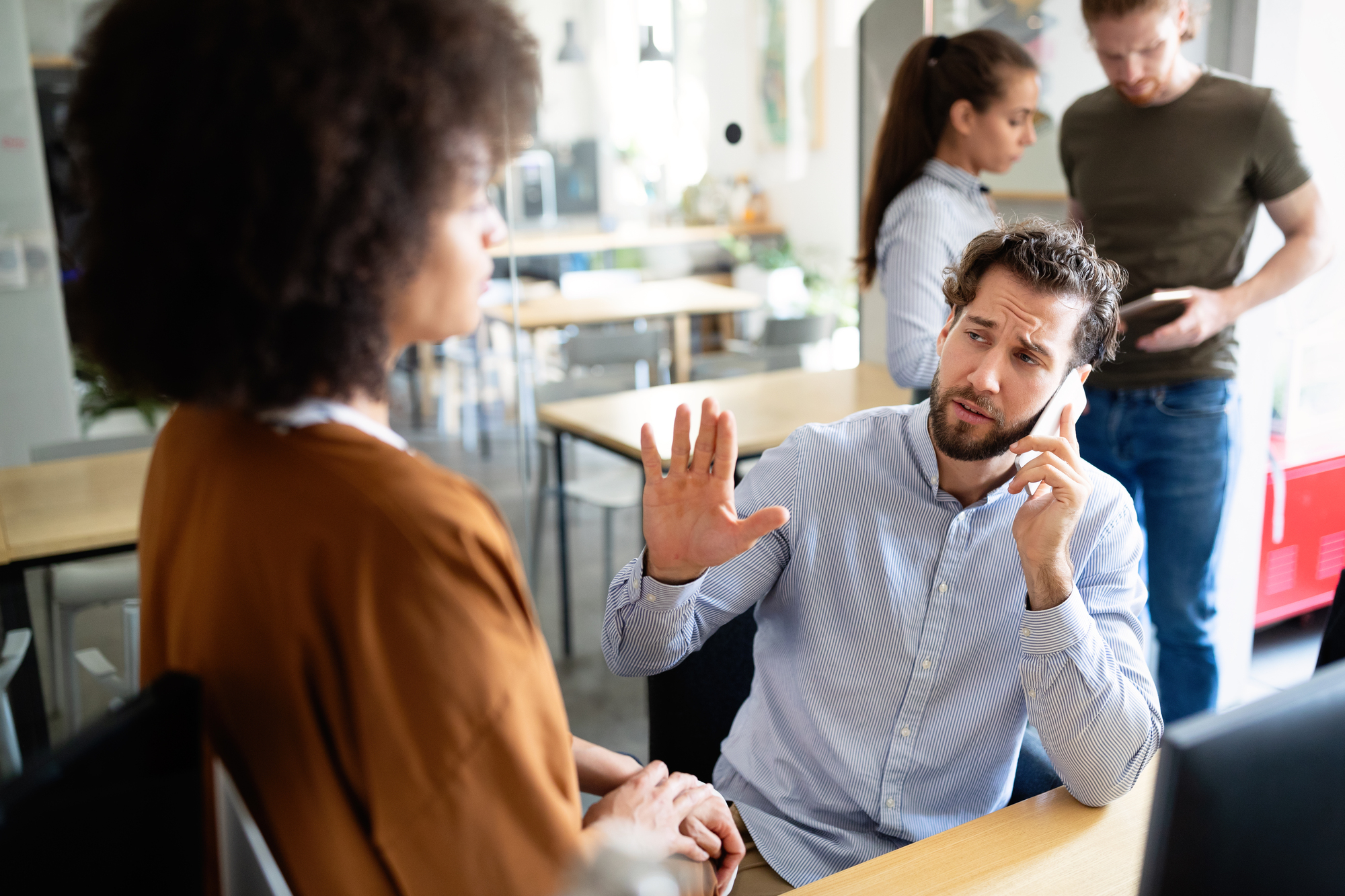 A man sitting at a desk holds up his hand while talking on the phone, appearing to be in a discussion with a woman standing in front of him. In the background, a woman on a laptop and a man on a smartphone are visible.