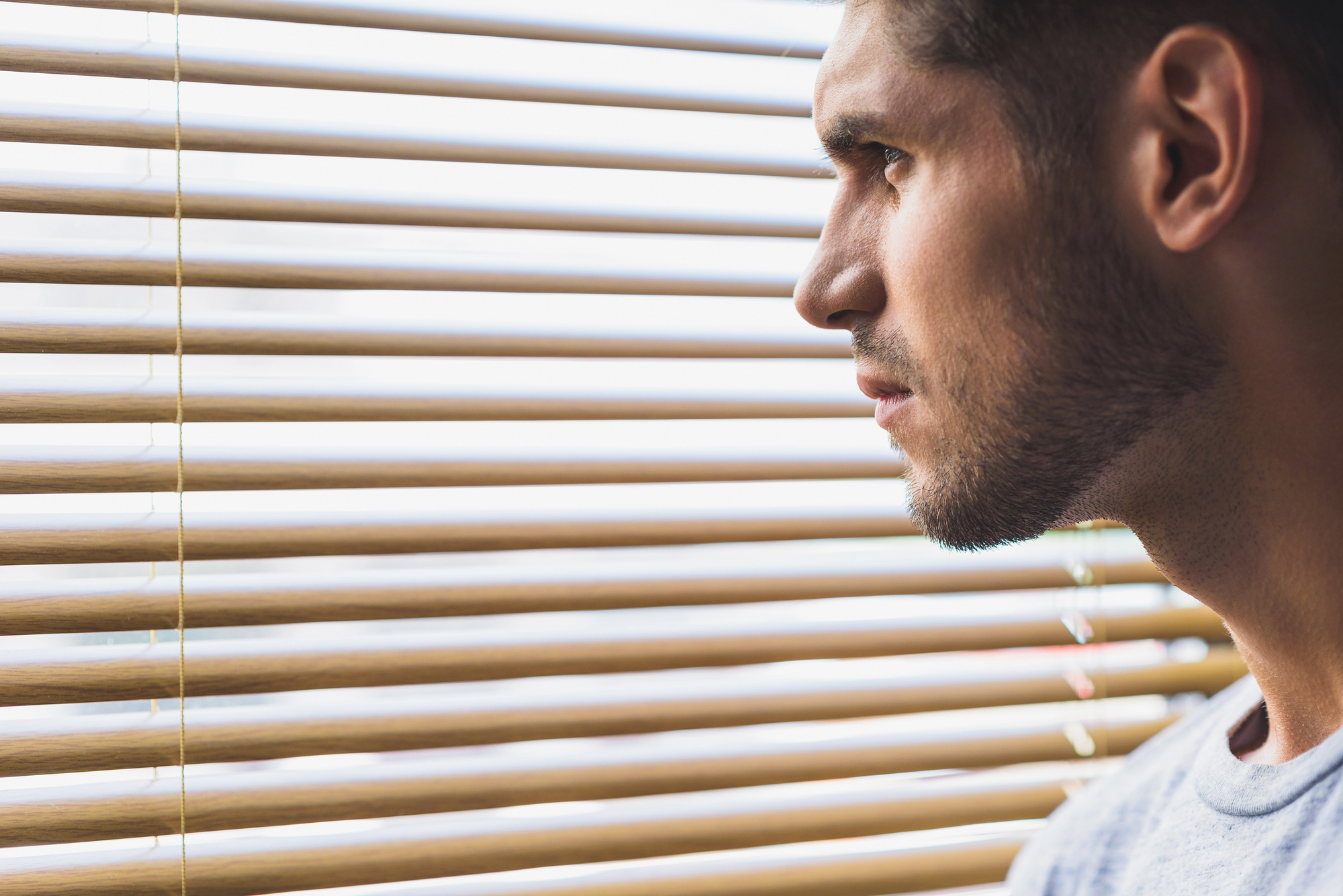 A man with short hair and a beard is looking pensively out through wooden blinds, with light streaming in, casting shadows on his face.