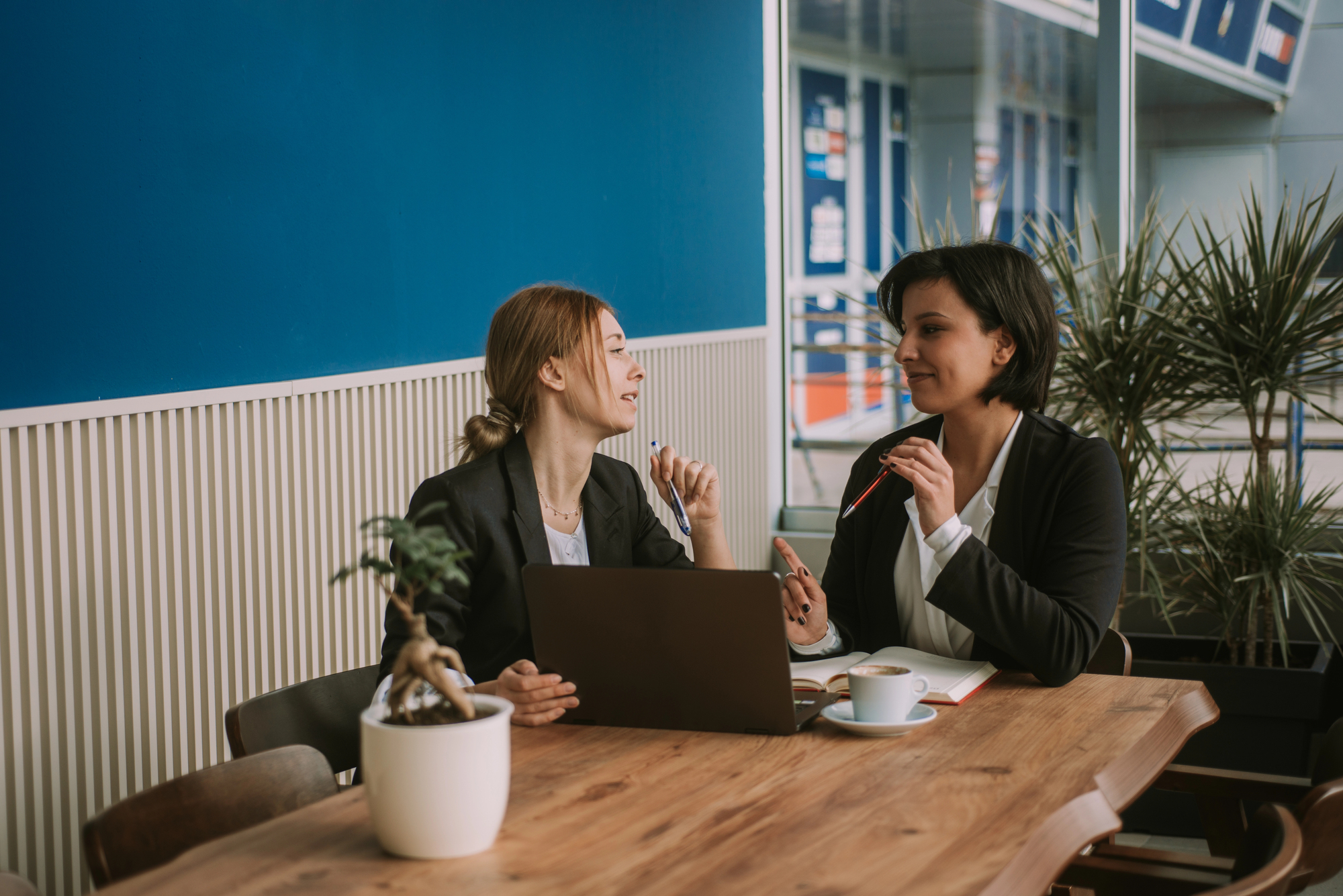 Two women in business attire sit at a wooden table with a laptop, engaged in conversation. One holds a pen and a cup of coffee is nearby. The background features indoor plants and a large blue wall.