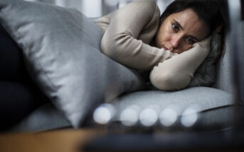 A person lying on a couch, wrapped in a blanket, with a somber expression. The individual is resting their head on a gray cushion and gazing thoughtfully forward. The foreground is slightly blurred, adding depth to the scene.