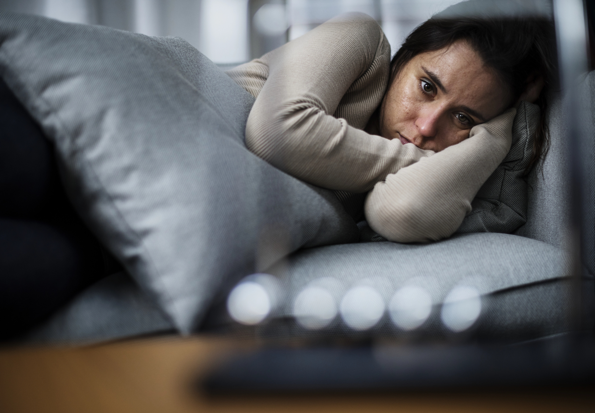 A person lying on a couch, wrapped in a blanket, with a somber expression. The individual is resting their head on a gray cushion and gazing thoughtfully forward. The foreground is slightly blurred, adding depth to the scene.