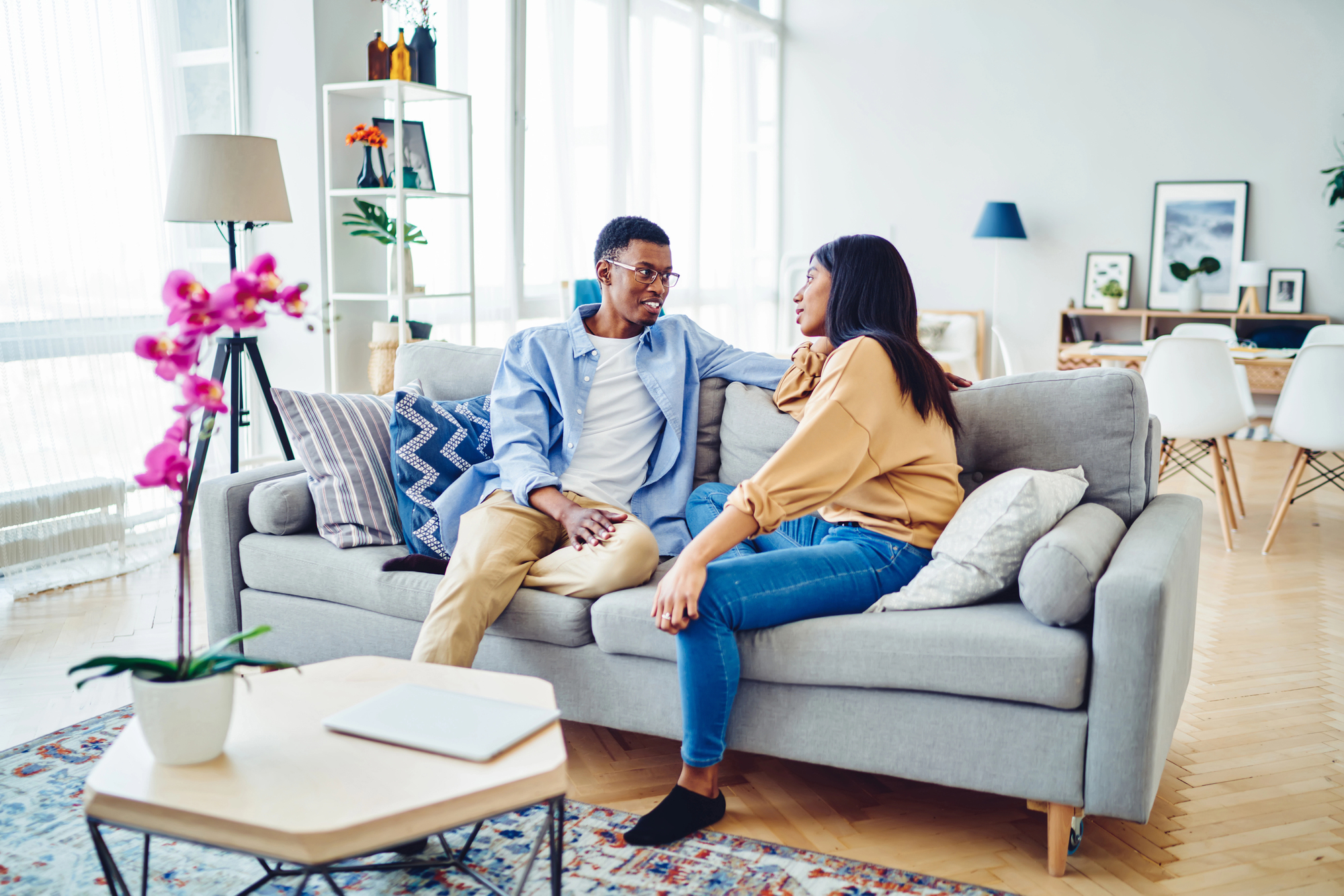 A couple sits on a gray sofa in a bright, modern living room. They are talking and smiling at each other. The room features decorative pillows, a lamp, plants, and a coffee table with a laptop. Large windows let in natural light.