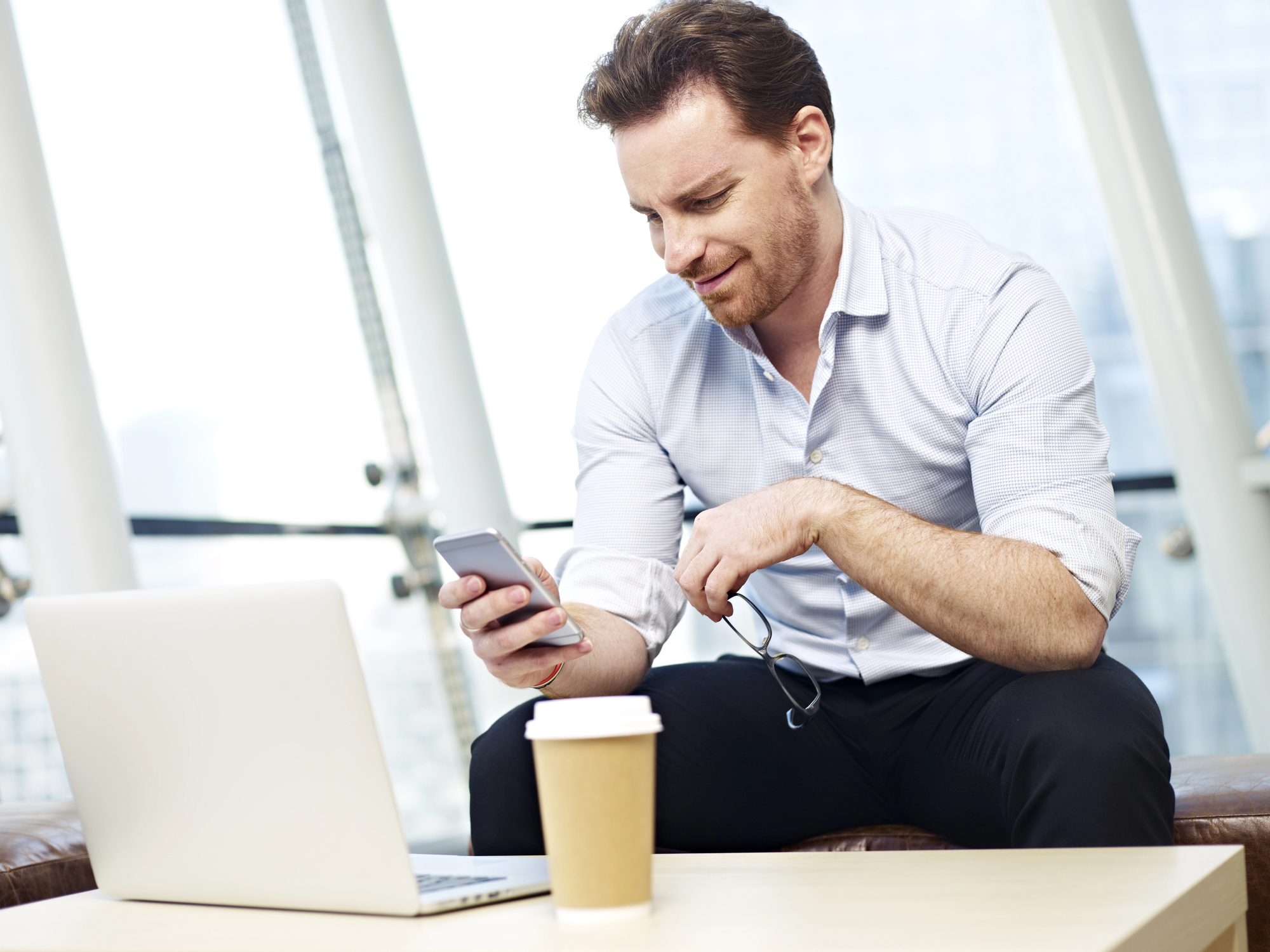 A man in a light shirt sits on a couch in a modern office space, holding a smartphone. He appears focused on the screen. A laptop and a disposable coffee cup are on the table in front of him. Sunlight streams in through large windows.