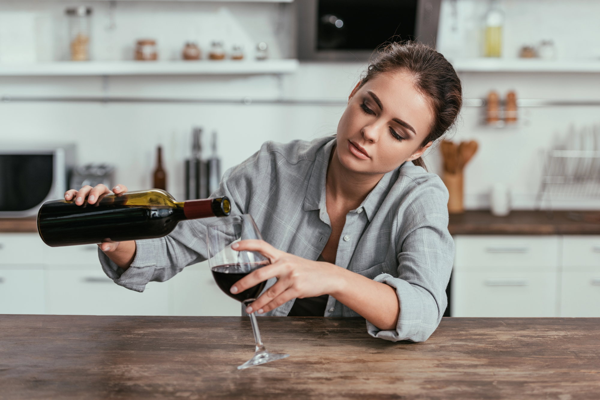 A woman in a gray shirt pours red wine into a glass in a modern kitchen, leaning on a wooden counter. Behind her, various kitchen items are visible, including bottles and utensils.