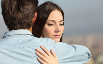 A woman with long brown hair appears thoughtful as she is embraced by a man with short brown hair. They both wear light blue shirts and are standing against a blurred outdoor background.