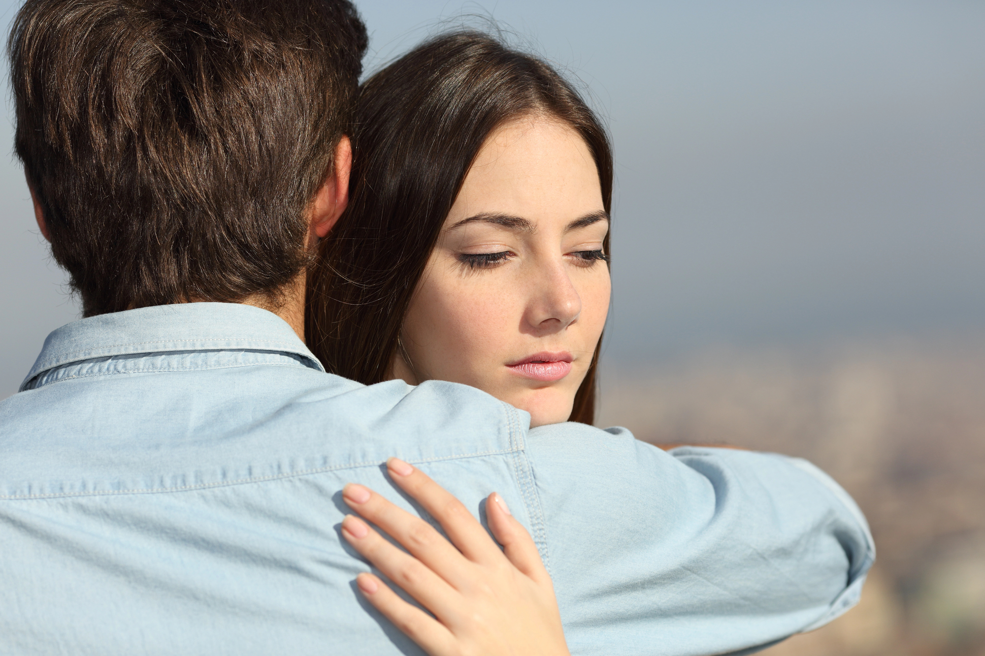 A woman with long brown hair appears thoughtful as she is embraced by a man with short brown hair. They both wear light blue shirts and are standing against a blurred outdoor background.