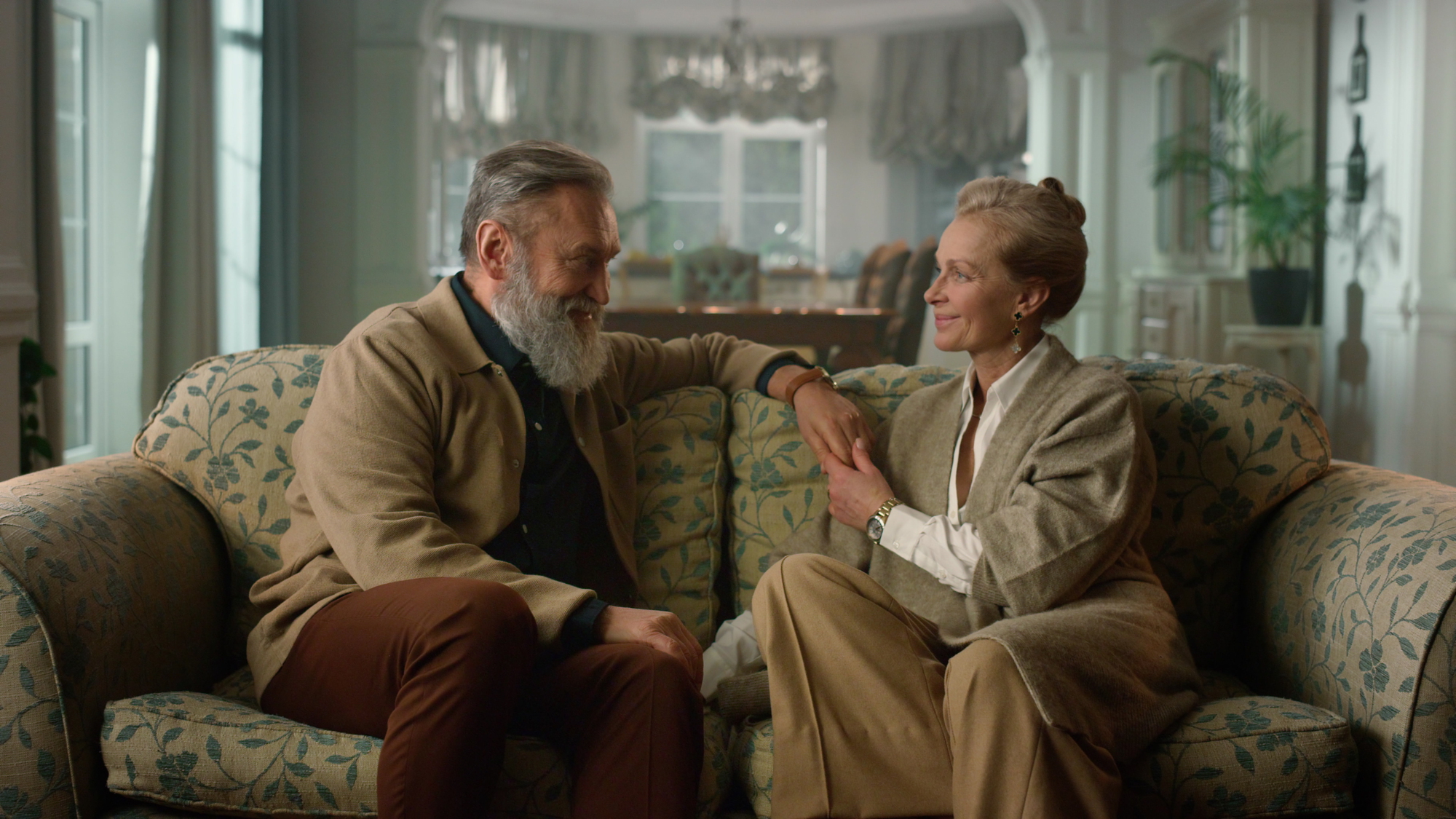 An elderly couple sits on a patterned sofa in a warmly lit living room. They are engaged in conversation, smiling at each other. Both are dressed in casual, beige-toned clothing, and the room has elegant decor with a dining area in the background.