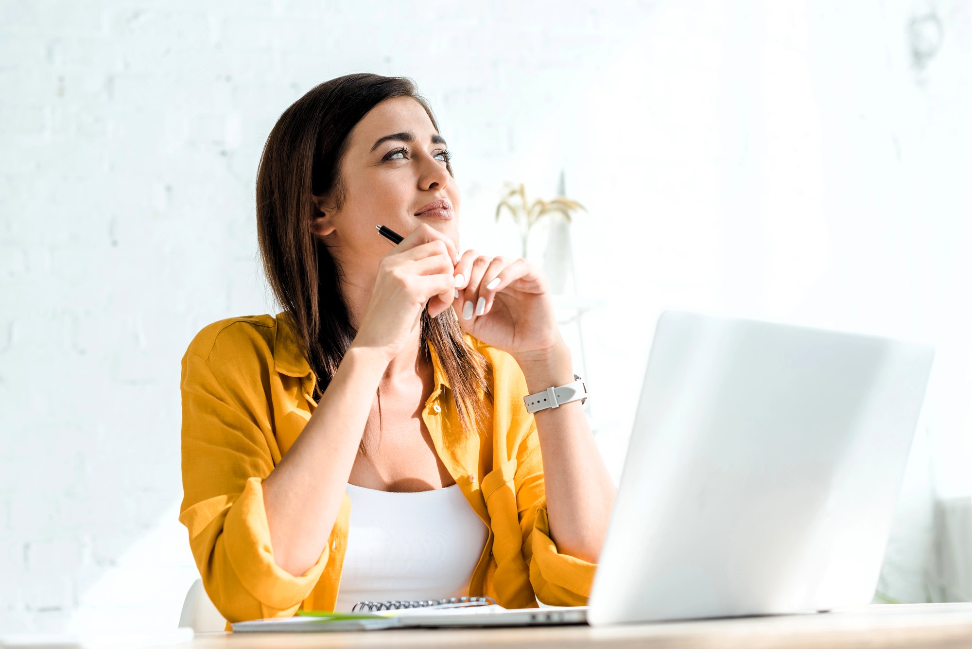 A woman in a yellow shirt sits at a desk with an open laptop. She is holding a pen to her chin, looking thoughtfully upwards. Sunlight illuminates the white wall behind her, giving the scene a bright, airy feel.