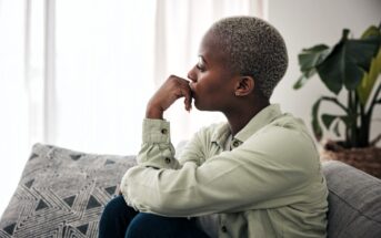 A person with short hair sits on a couch, looking thoughtfully out a window. They wear a light-colored shirt and have one hand resting under their chin. A patterned cushion and a large plant are visible in the background.