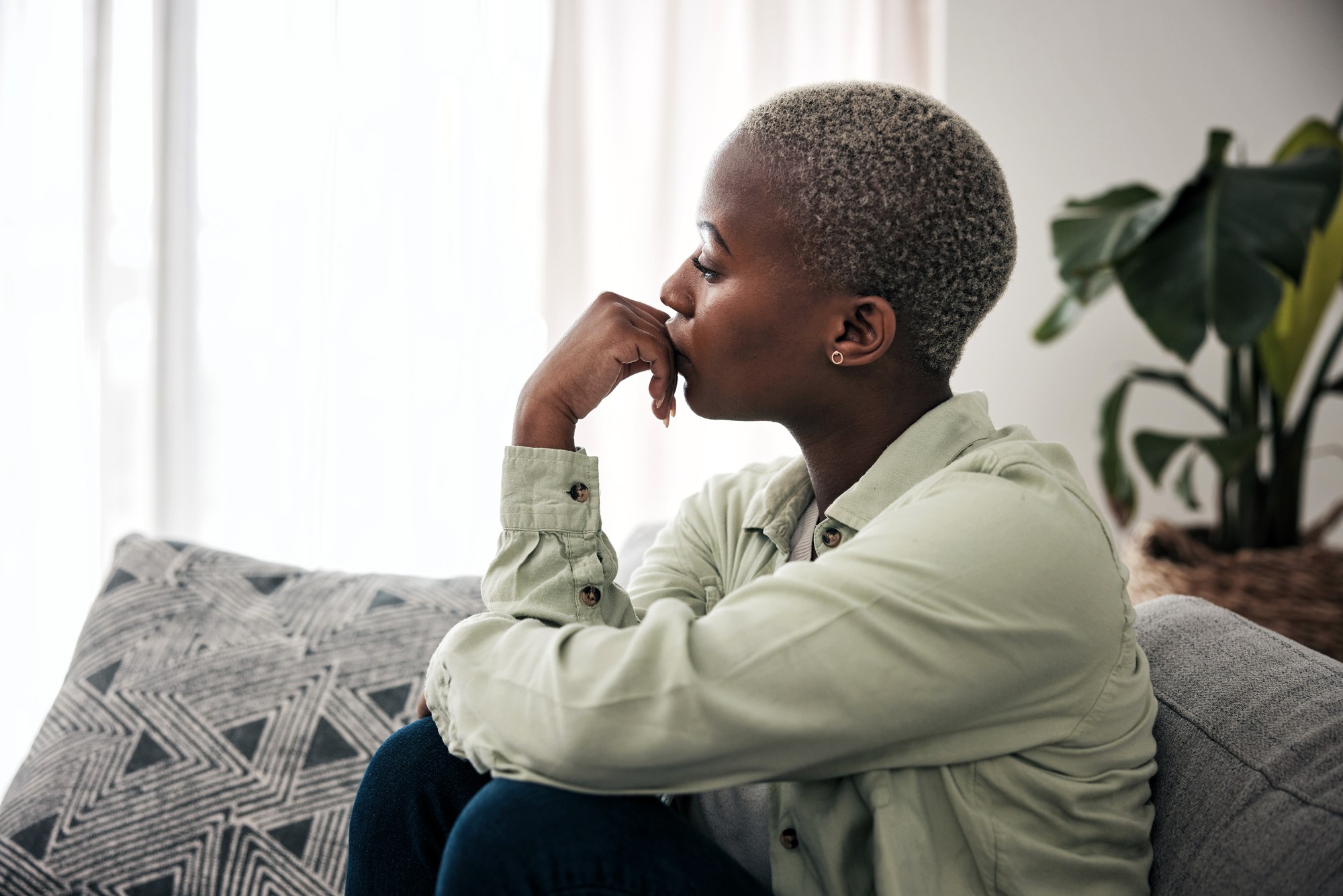 A person with short hair sits on a couch, looking thoughtfully out a window. They wear a light-colored shirt and have one hand resting under their chin. A patterned cushion and a large plant are visible in the background.