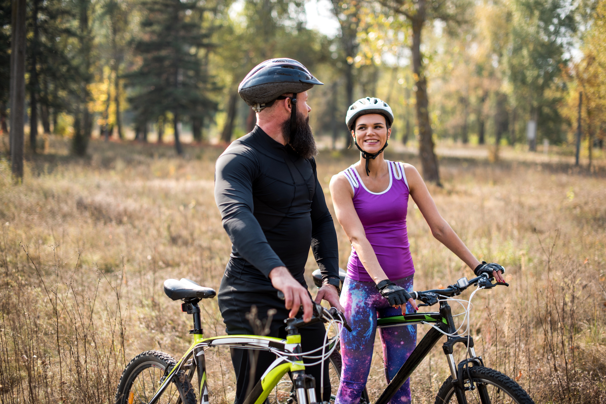 A man and a woman wearing helmets stand with their bicycles in a wooded area during autumn. The woman wears a purple tank top and patterned leggings, while the man wears a black long-sleeve shirt. They are smiling at each other.