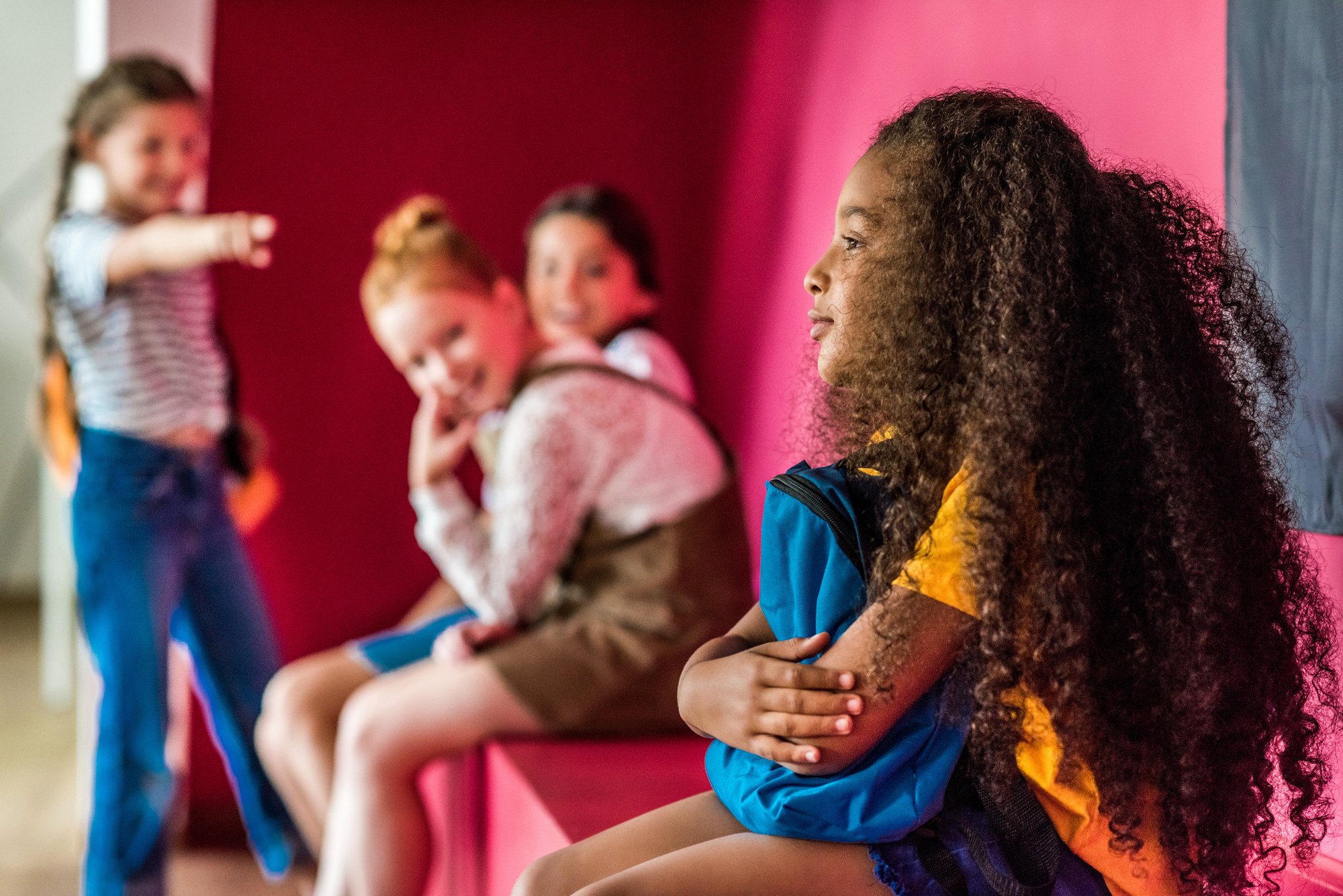 A child with long curly hair sits alone, looking away, while three children in the background sit together, with one pointing and smiling. The setting has vibrant red walls.