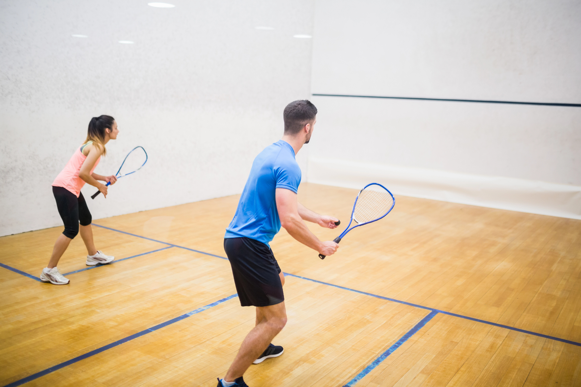 Two people playing squash on a wooden court. The man in blue prepares to hit the ball, while the woman in pink stands ready behind him. White walls with court markings are visible in the background. Both hold squash rackets.