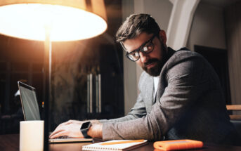 A man with glasses and a beard sits at a table working on a laptop. He is wearing a gray blazer and looking over his shoulder. The setting is indoors with a warm lamp, a notebook, and an orange external hard drive on the table.