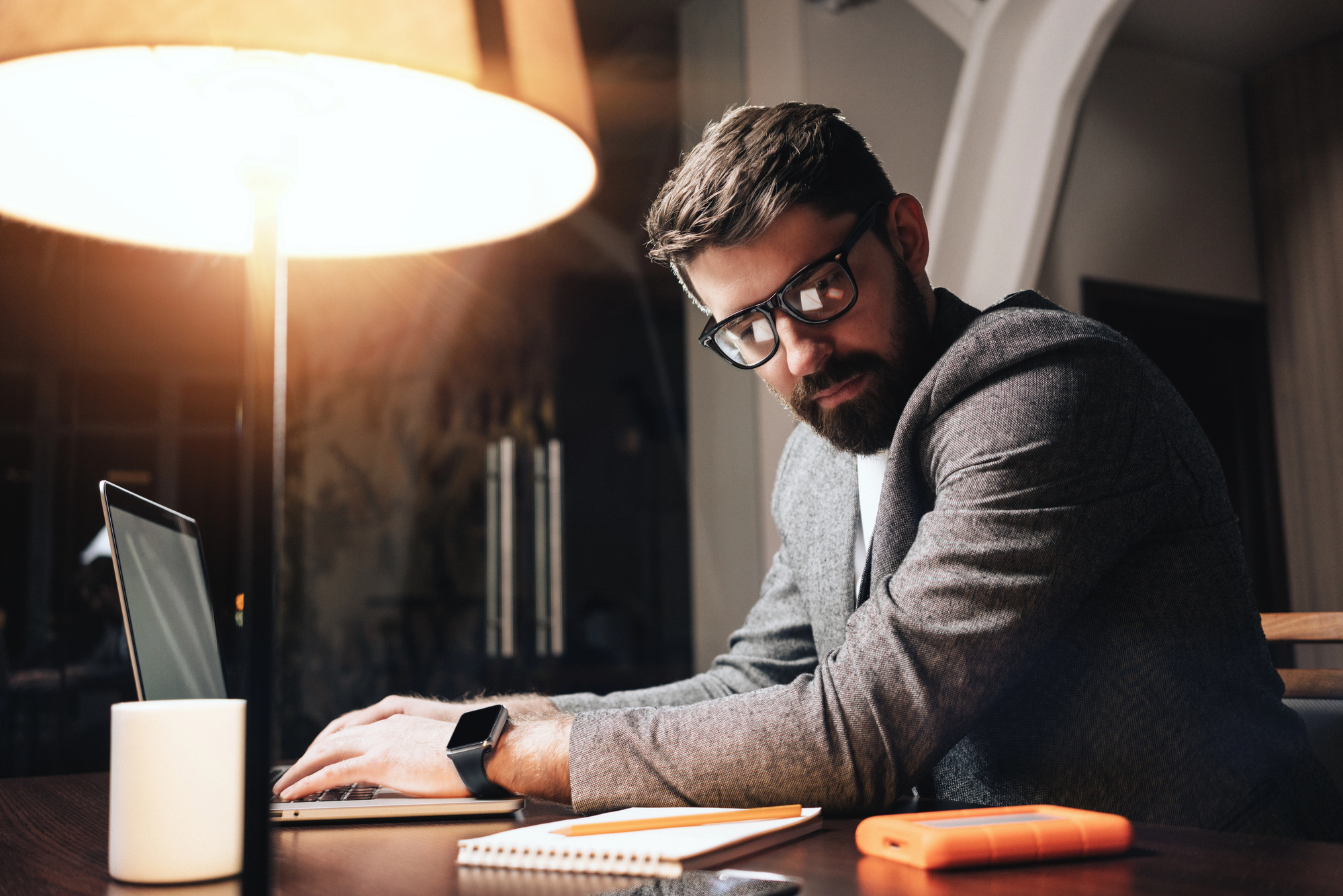 A man with glasses and a beard sits at a table working on a laptop. He is wearing a gray blazer and looking over his shoulder. The setting is indoors with a warm lamp, a notebook, and an orange external hard drive on the table.