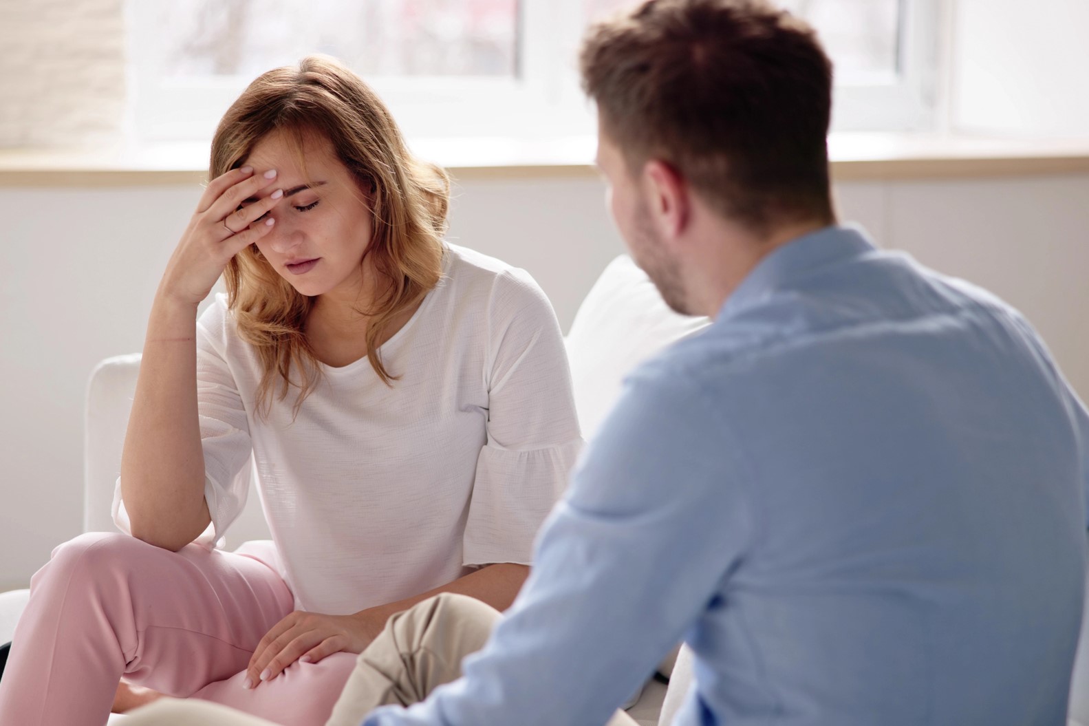 A woman sitting on a couch with her hand on her forehead, looking stressed or upset. She is wearing a white shirt and pink pants. A man is sitting next to her, turned towards her, appearing to listen attentively.