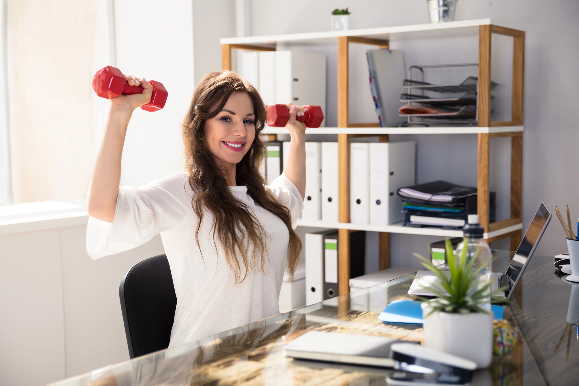 A woman sitting at a desk in an office, smiling, while lifting red dumbbells. The desk has a laptop, books, and a potted plant. Shelves with binders and files are in the background. Natural light from a window brightens the space.