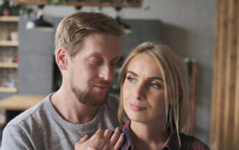 A man and woman stand close together in a cozy room. The man, with short blonde hair and a beard, looks at the woman fondly. The woman, with long blonde hair, looks ahead with a slight smile. Both wear casual clothing, creating a relaxed atmosphere.