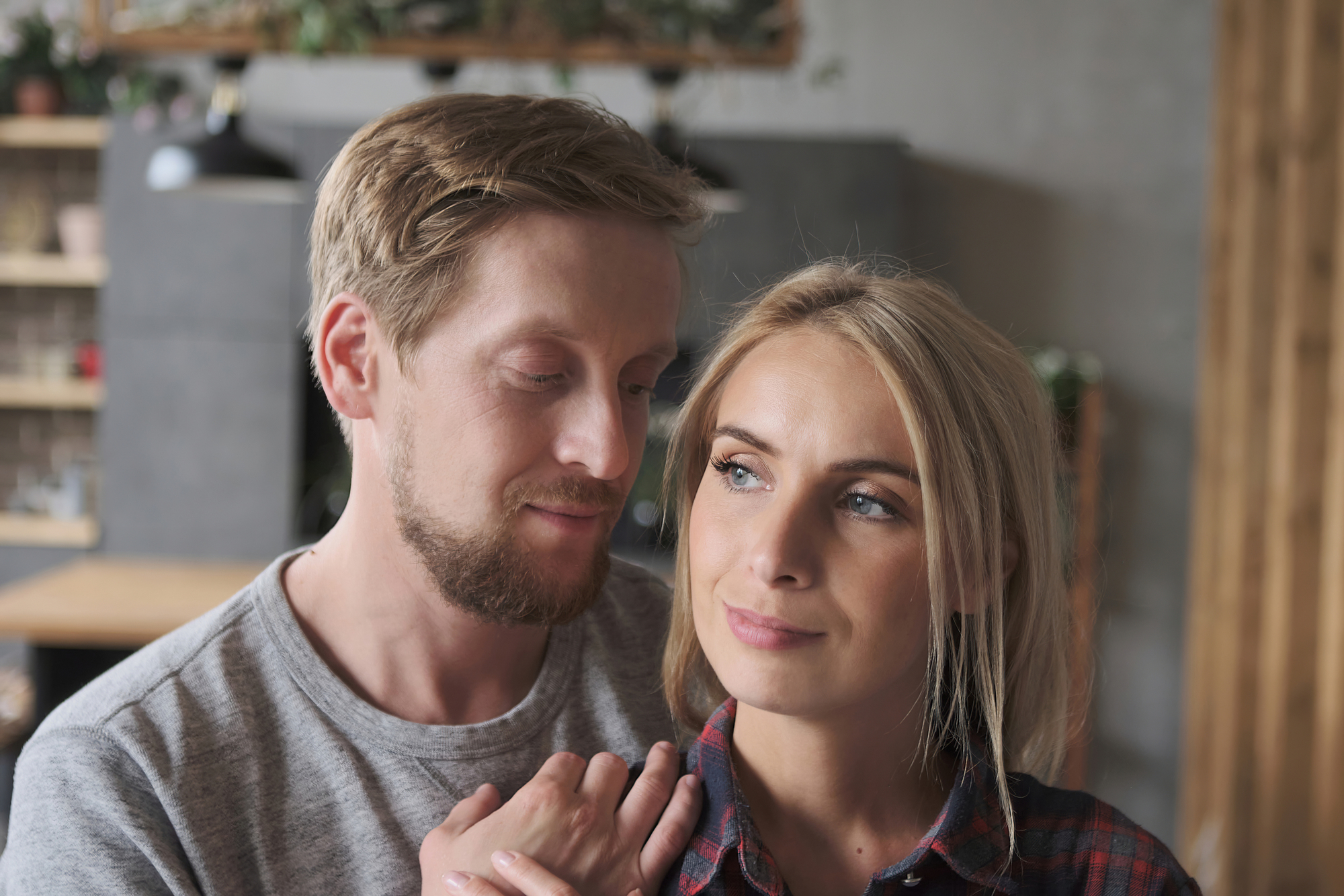 A man and woman stand close together in a cozy room. The man, with short blonde hair and a beard, looks at the woman fondly. The woman, with long blonde hair, looks ahead with a slight smile. Both wear casual clothing, creating a relaxed atmosphere.