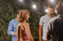 A woman in a red and white striped dress smiles while talking to three men at an outdoor gathering. String lights hang above, and greenery is visible in the background.