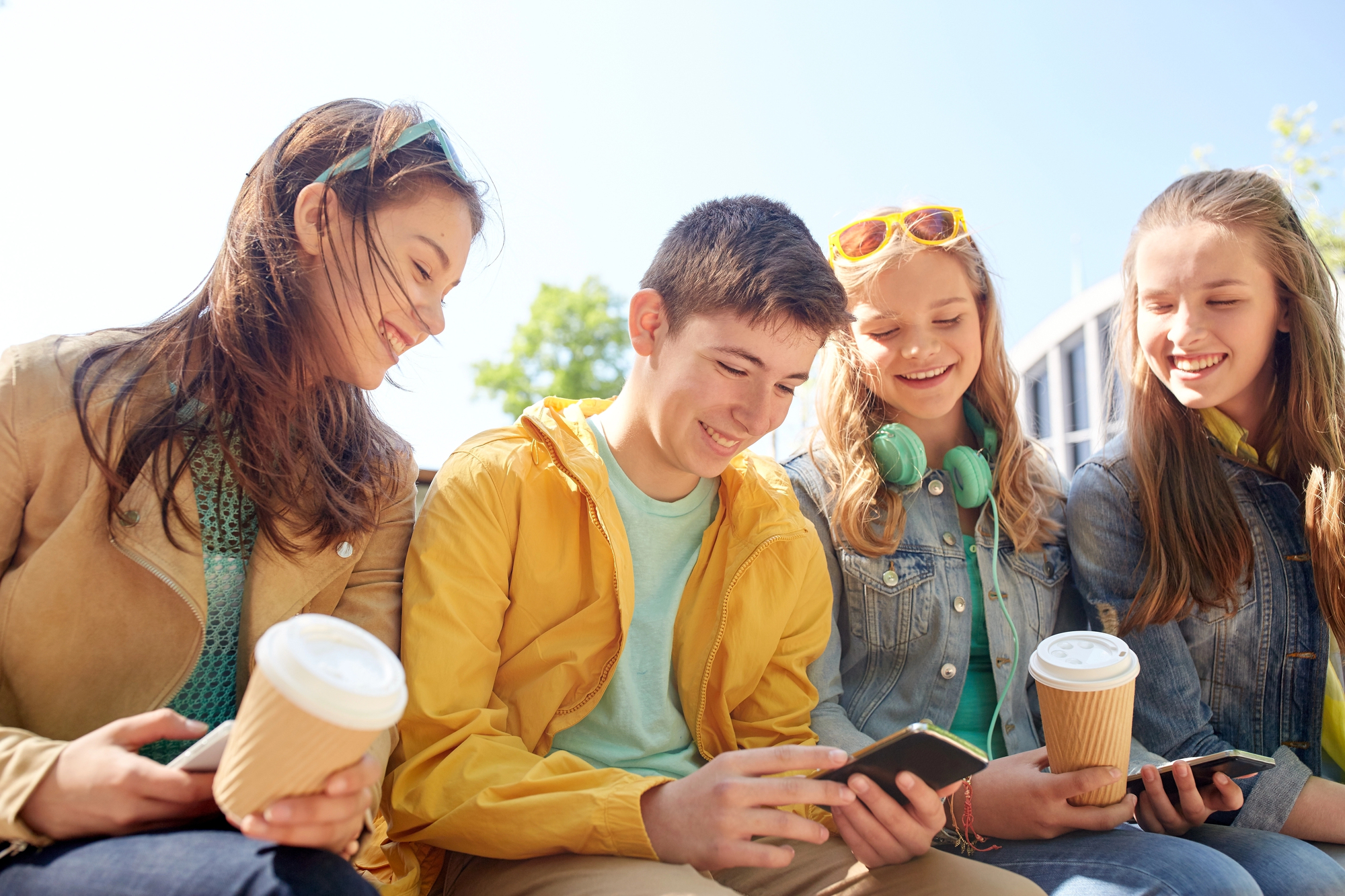 Four teenagers sitting outside, smiling and looking at a smartphone. They are casually dressed; two hold takeout coffee cups, and one wears headphones, while another has sunglasses on their head. It's a sunny day with a clear sky.