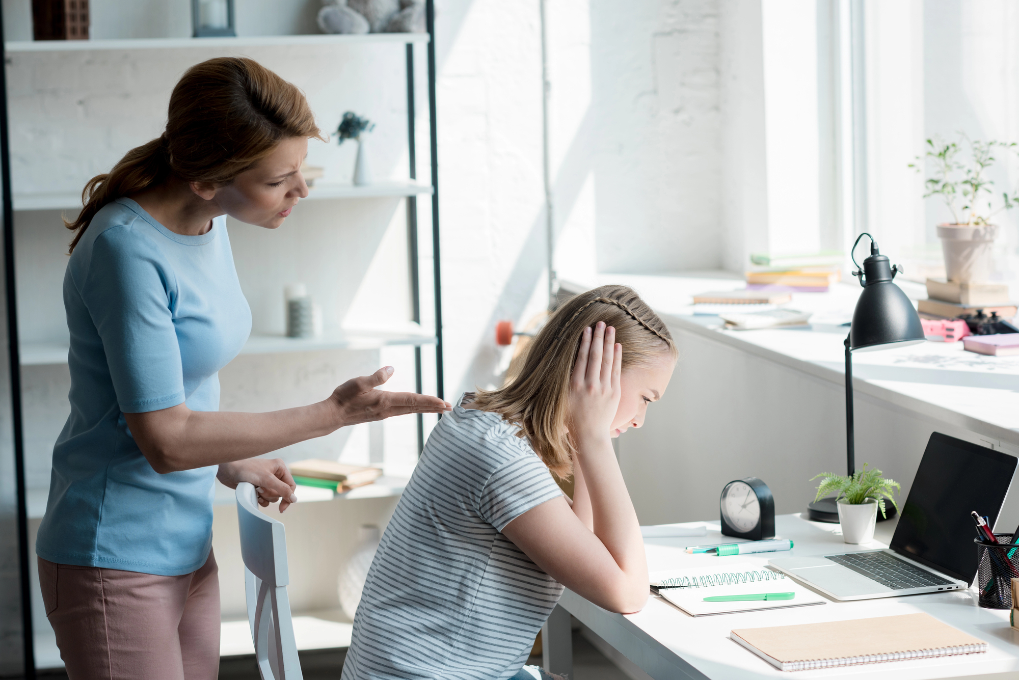 A woman is talking to a girl who covers her ears and looks upset, sitting at a desk with stationery, a laptop, and a clock. The scene is lit by natural light from a large window, and the room has light-colored walls and shelves.