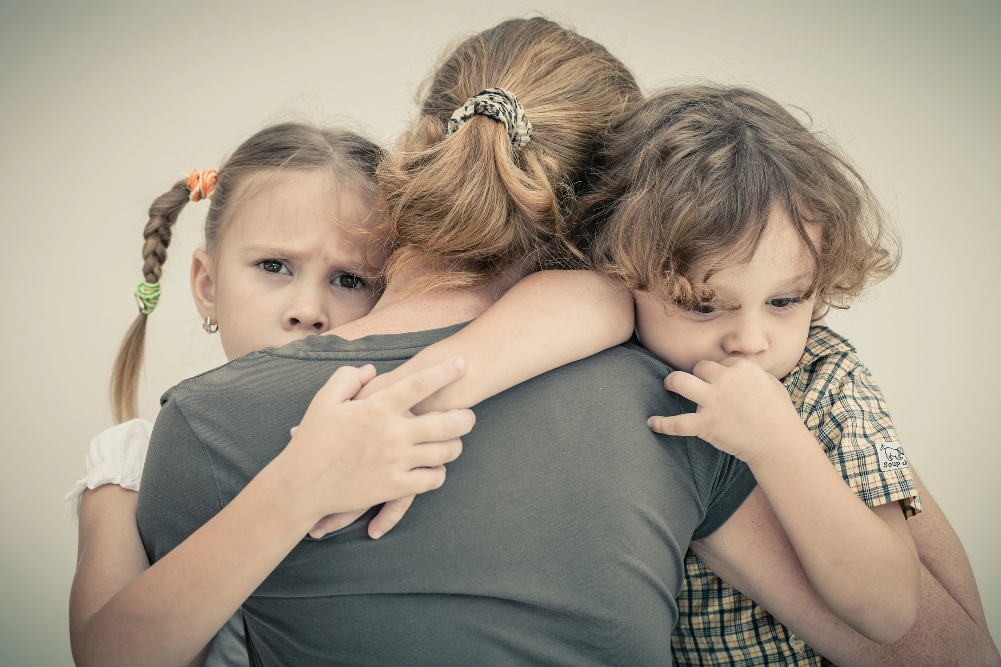 A woman with her back to the camera hugs two children. The girl on the left looks concerned, with braids and a white shirt. The boy on the right is holding his hand to his mouth, wearing a checkered shirt. They appear comforted by the embrace.