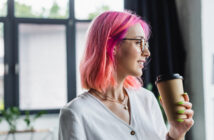 A person with pink hair and glasses holds a coffee cup and smiles, standing indoors against large windows. They wear a white shirt and have neon green nails.