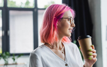 A person with pink hair and glasses holds a coffee cup and smiles, standing indoors against large windows. They wear a white shirt and have neon green nails.