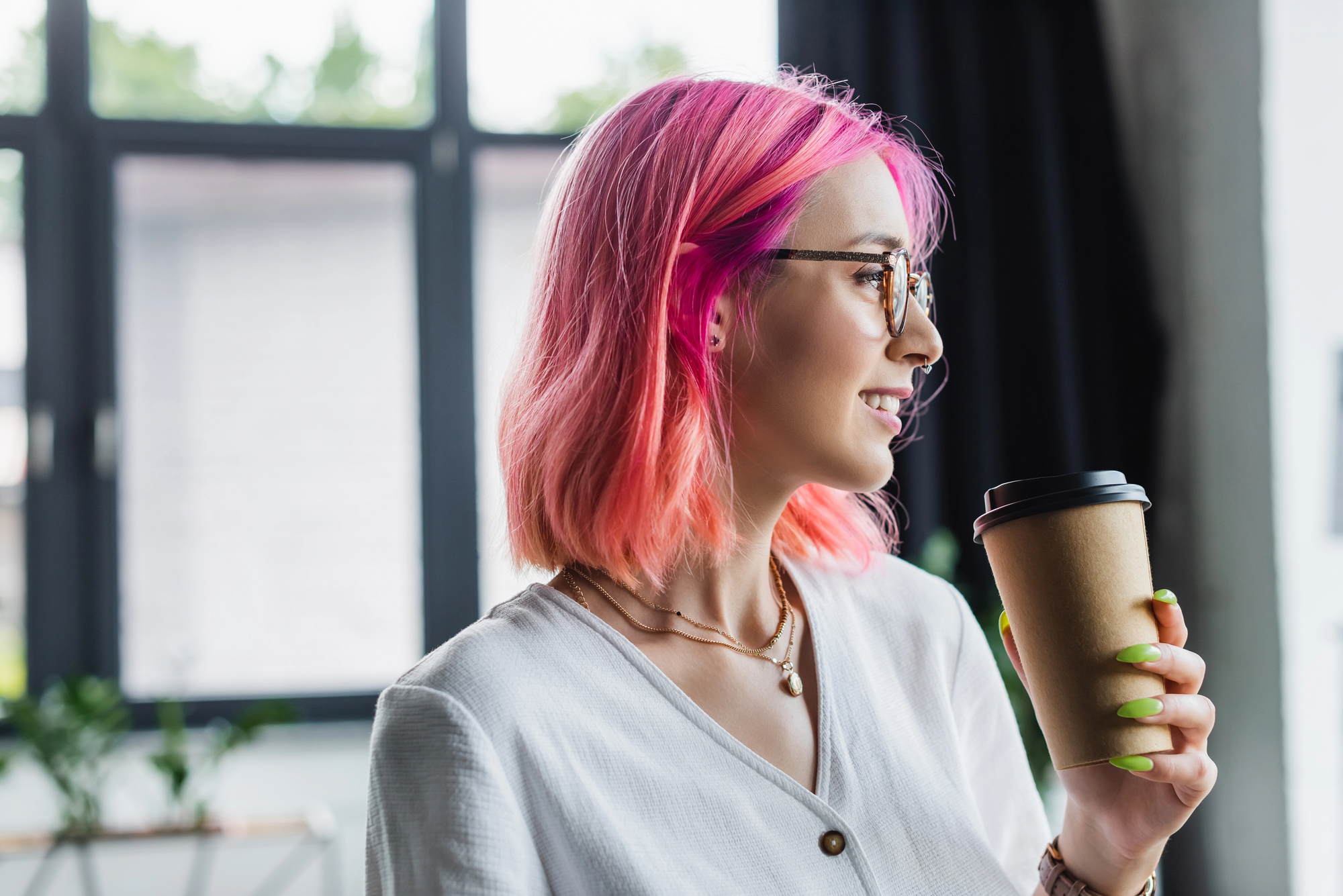 A person with pink hair and glasses holds a coffee cup and smiles, standing indoors against large windows. They wear a white shirt and have neon green nails.