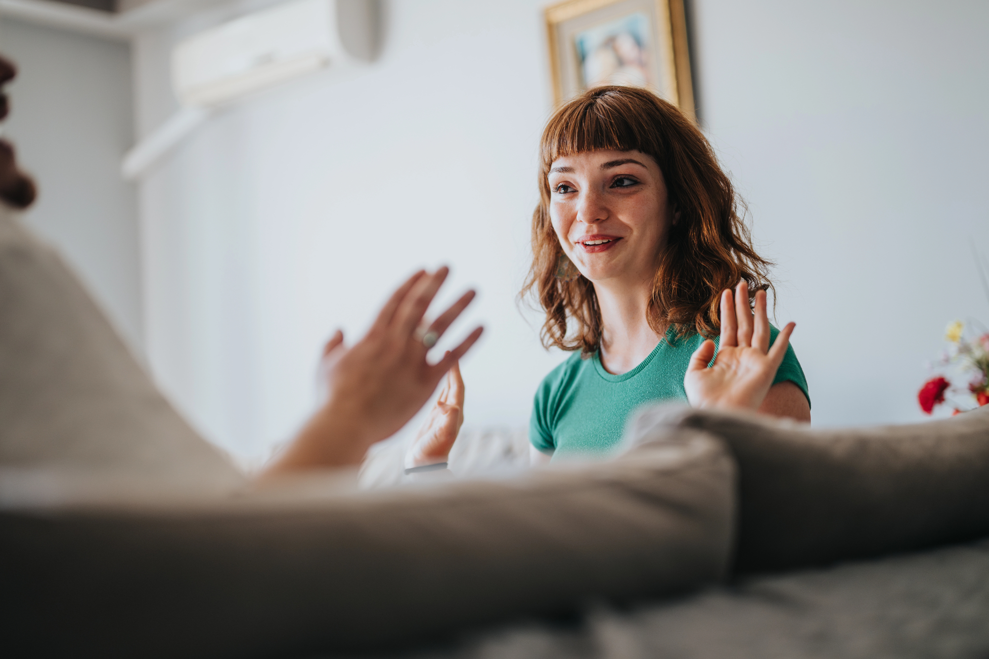 A woman with red hair, wearing a green shirt, sits on a couch engaged in an animated conversation, gesturing with her hands. A framed picture and air conditioner are visible in the background.