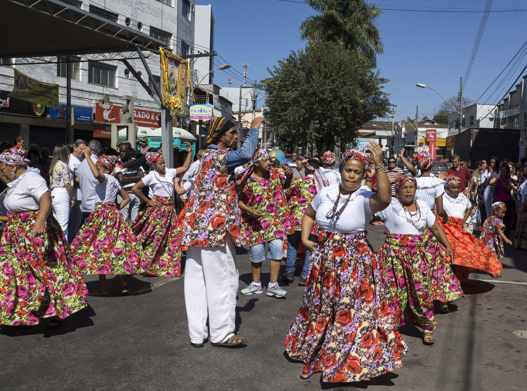 A vibrant street scene with a group of dancers in colorful floral skirts and headbands. They are performing energetically, surrounded by onlookers in casual summer attire. The background features buildings and a clear blue sky.