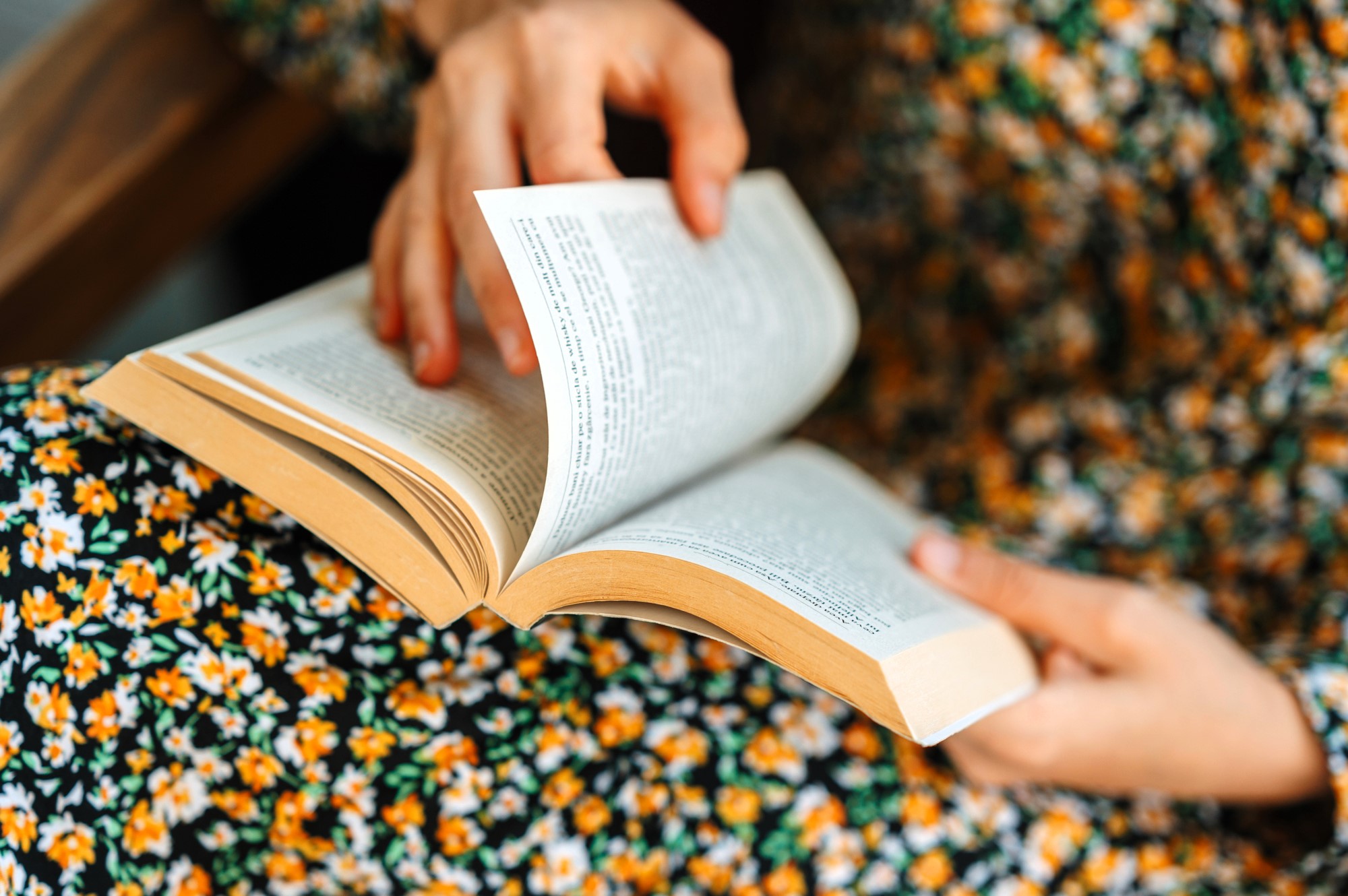 A person wearing a floral-patterned dress is reading a book, holding it open with both hands. The focus is on the hands and the book, with pages gently being turned. The background is blurred, emphasizing the book and dress pattern.