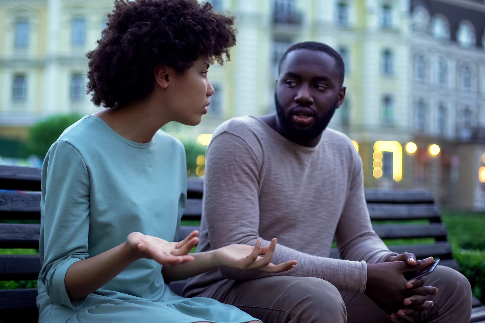 A man and a woman sitting on a bench outdoors. The woman is gesturing with her hands while talking, and the man is looking at her, holding a smartphone. Buildings and lights are blurred in the background.