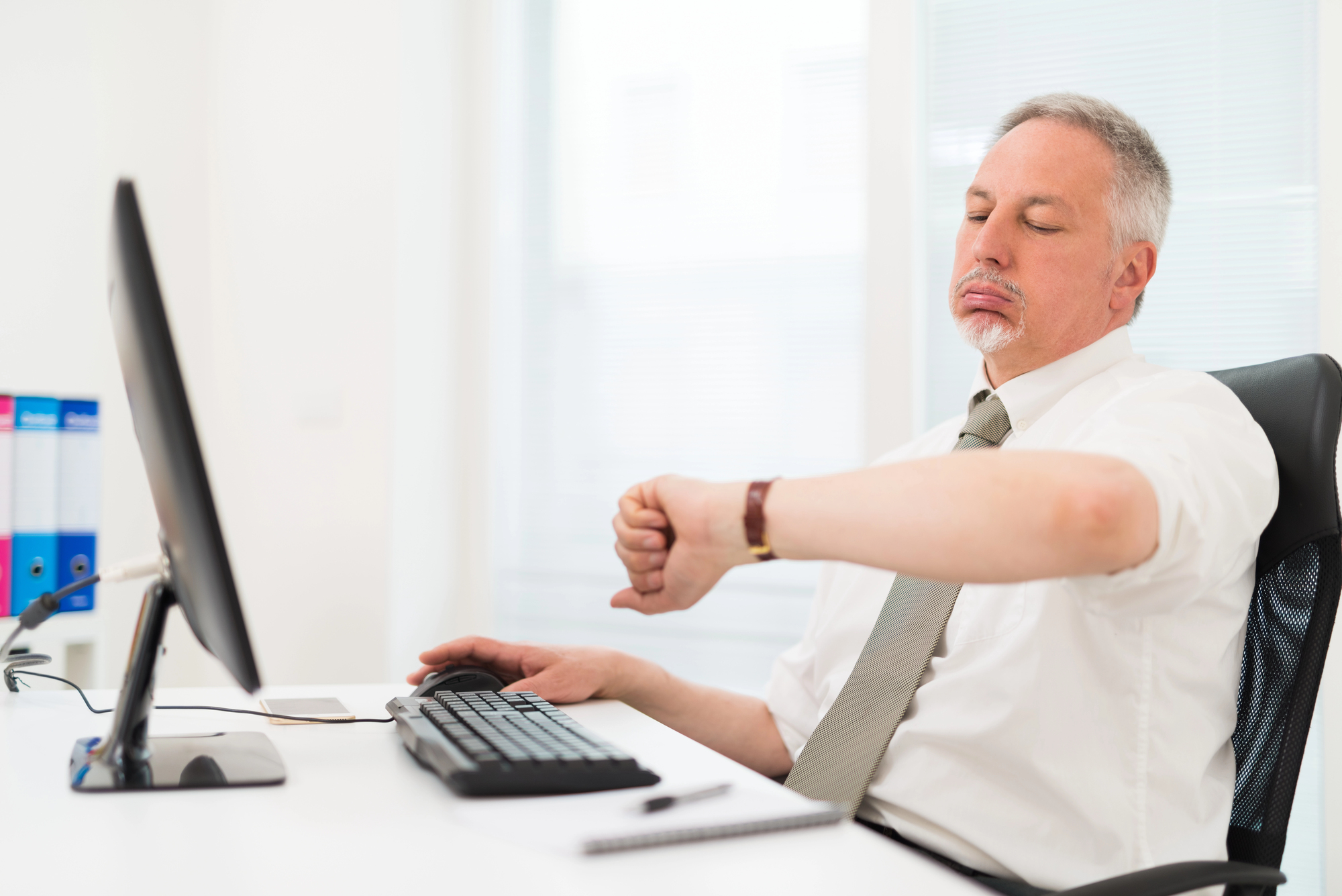 A middle-aged man in a white shirt and tie sits at a desk in an office, looking at his watch with an impatient expression. He is at a computer, with folders and a notepad nearby. Bright natural light comes through a window.